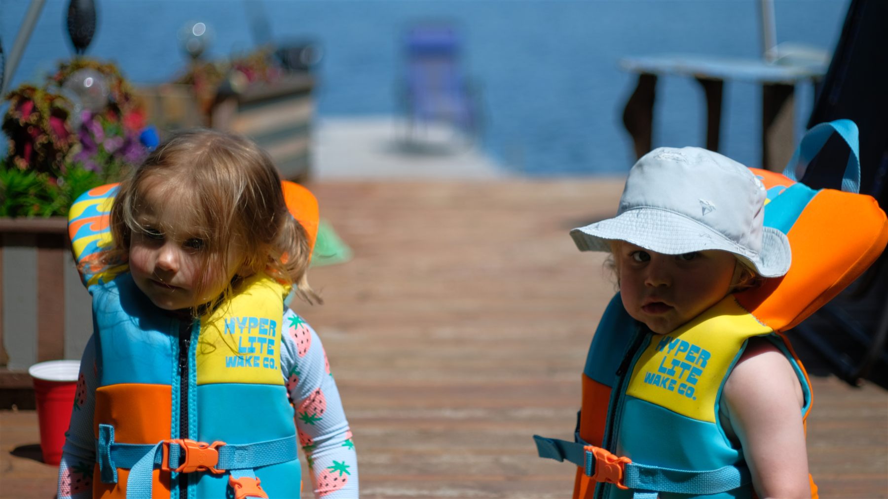 Two toddlers in life jackets standing on a dock by the lake